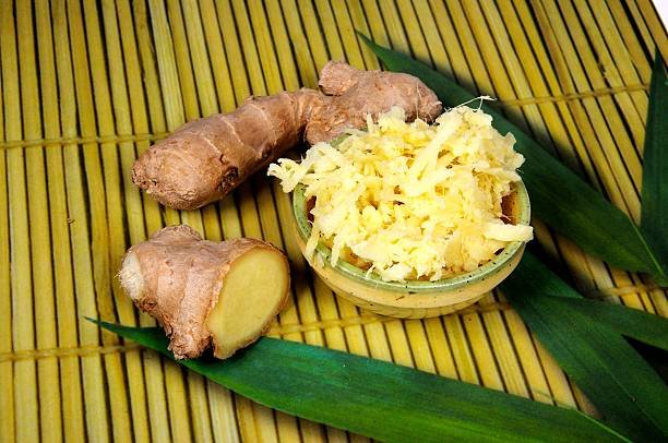Two pieces of root ginger with a dish of grated ginger against a bamboo background.